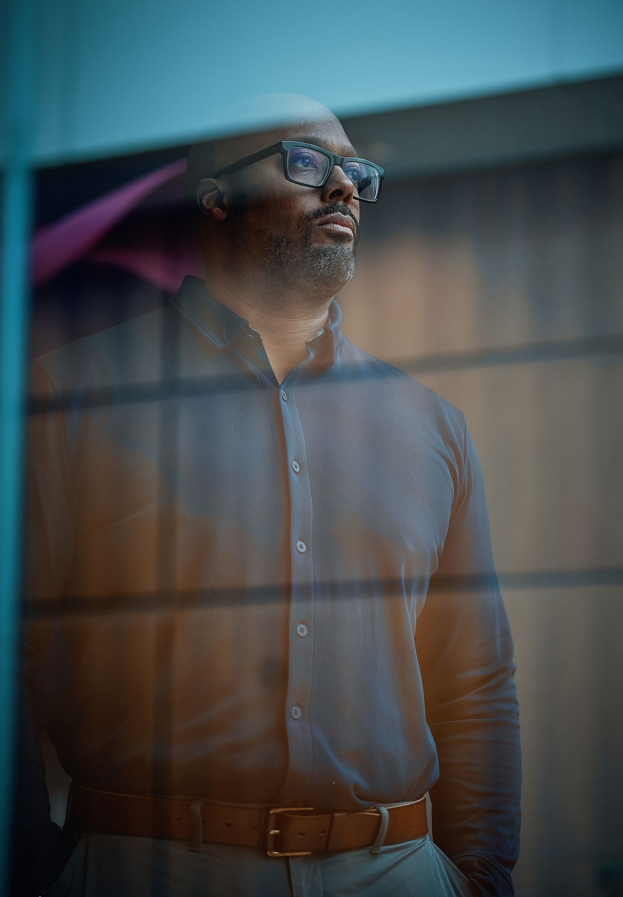 Man with glasses gazes out of a window, wearing a dark shirt and light brown pants, with a thoughtful expression.