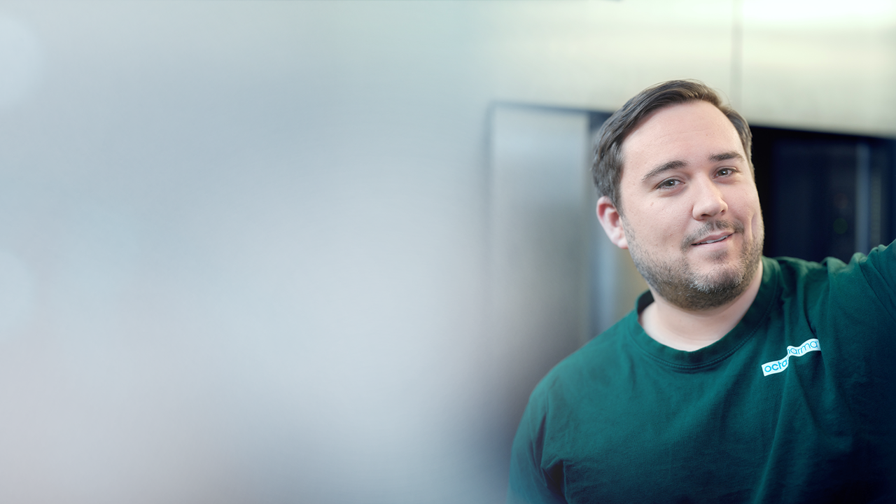 An Octapharma production employee in a green shirt stands indoors with a blurred background, looking towards the camera, while reaching out with one arm.