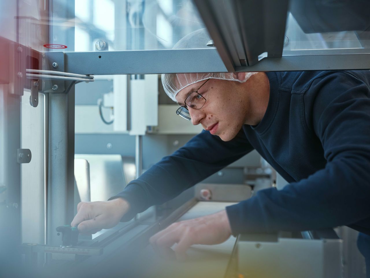 A man wearing glasses and a hairnet works intently on machinery in a bright industrial setting.