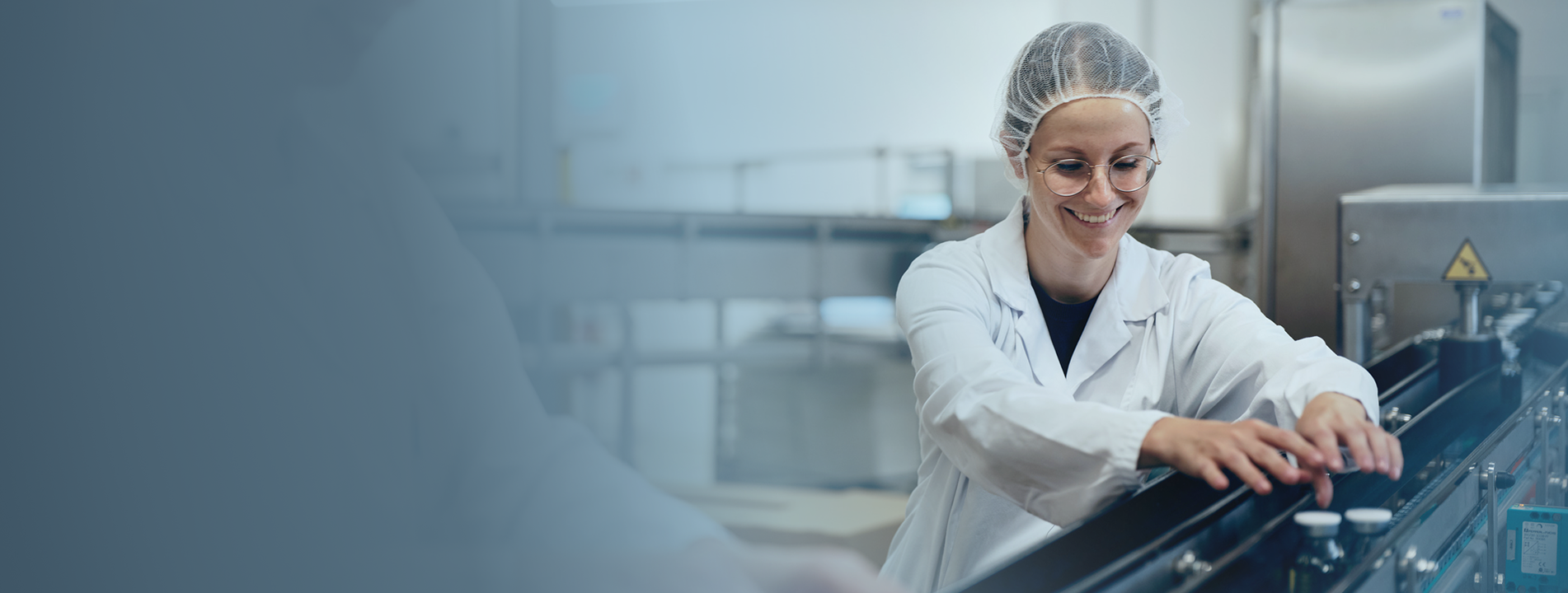 Smiling employee in lab coat and hairnet operates a conveyor belt at an Octapharma production site.