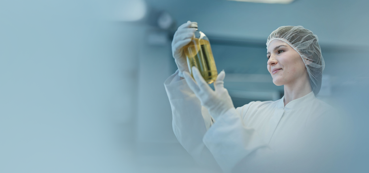 Employee in protective clothing examining a glass container with liquid at an Octapharma production facility.