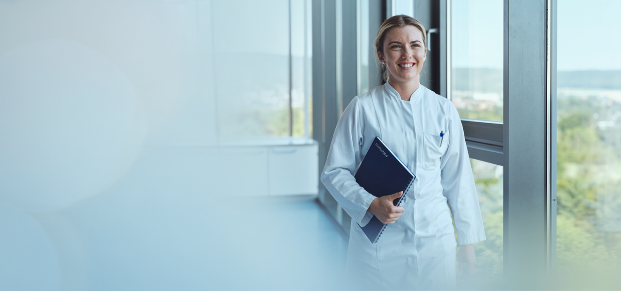 Smiling Octapharma employee in a white lab coat standing by a window, holding a notebook.