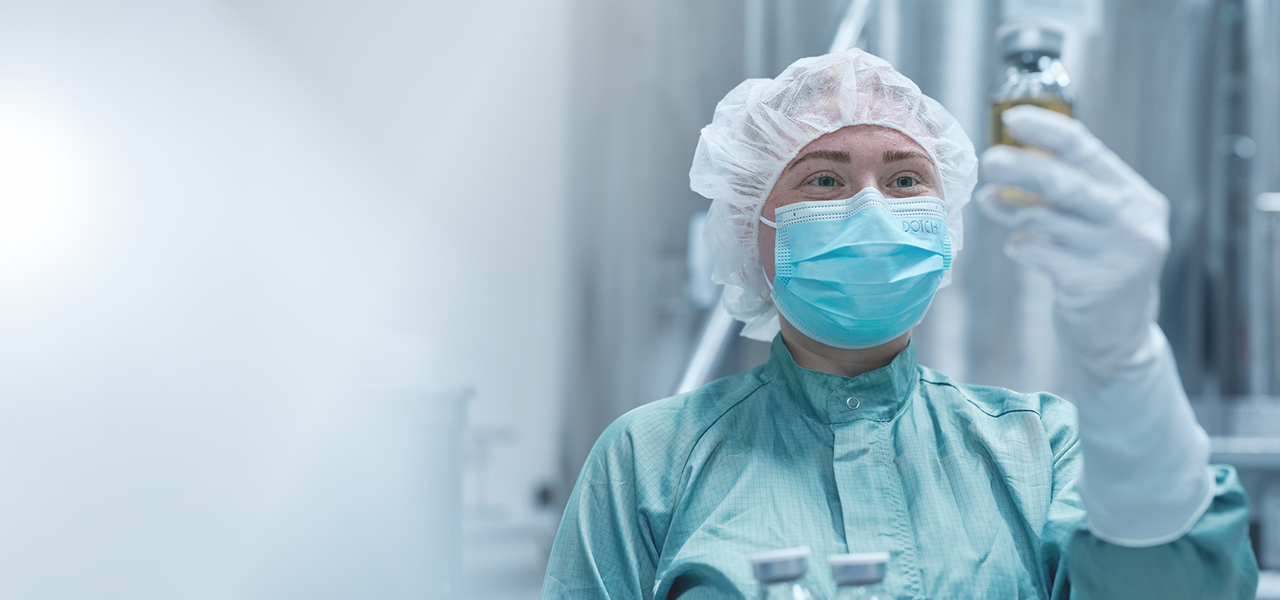 Octapharma-Mitarbeiterin in Schutzkleidung und Maske bei der Inspektion von Glasfläschchen in einer Produktionsstätte.