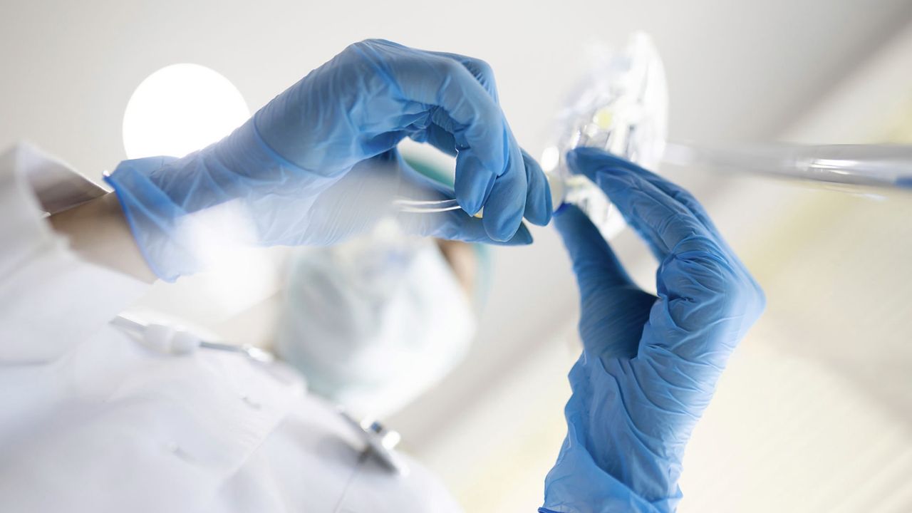Healthcare professional wearing gloves and a mask examines an IV bag in a bright clinical environment.