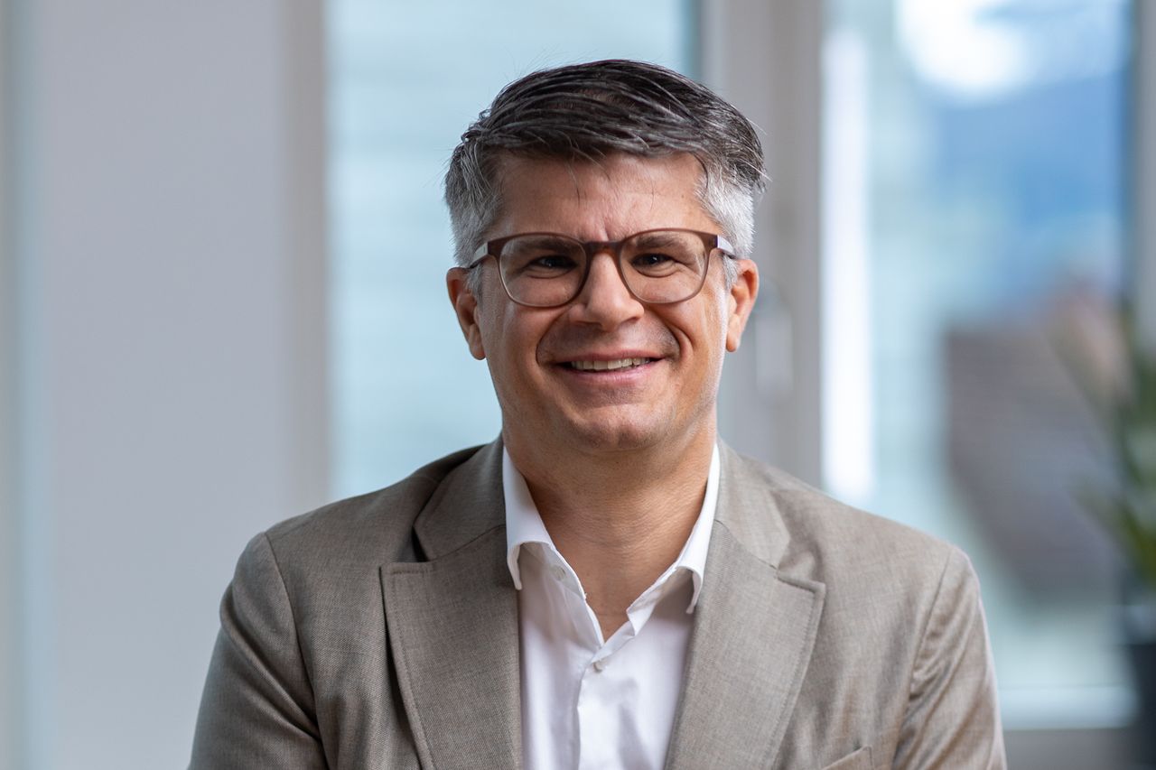 Roger Mächler, Chief Financial Officer, wearing a beige suit and white shirt, sitting indoors with a blurred window background.