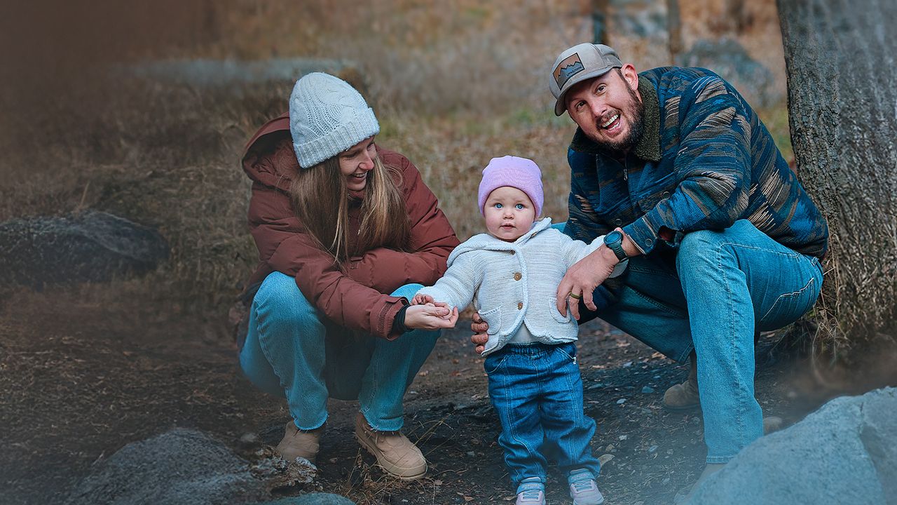 A smiling couple kneels beside a toddler in a winter scene, all wearing warm clothing. The toddler stands between them, holding their hands.