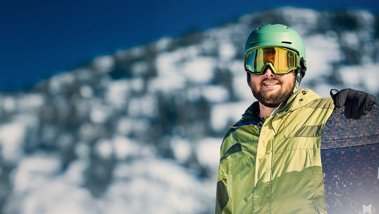 A snowboarder in a green jacket and helmet stands with a snowboard against a snowy mountain backdrop on a sunny day.
