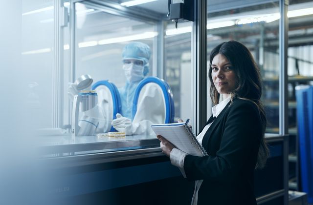 Woman in a suit holds a notebook, standing by a lab with a technician in a protective suit behind glass, working with blue gloves.