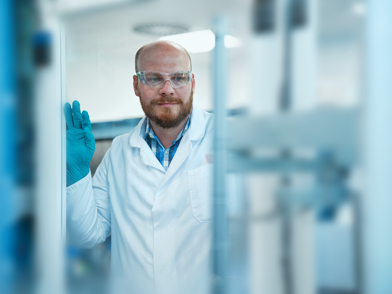 A scientist in a lab coat and safety glasses is working in a laboratory, holding up a gloved hand while near scientific equipment.