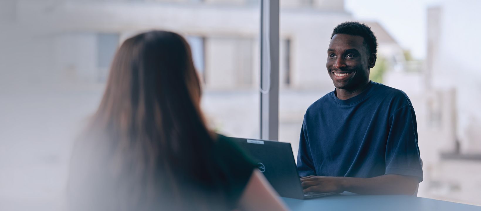 Two people sit at a table in an office setting, one smiling with a laptop, in conversation.