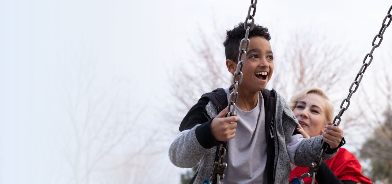 A joyful child is swinging on a swing, holding the chains, with a smiling woman gently pushing him. Trees are visible in the background.