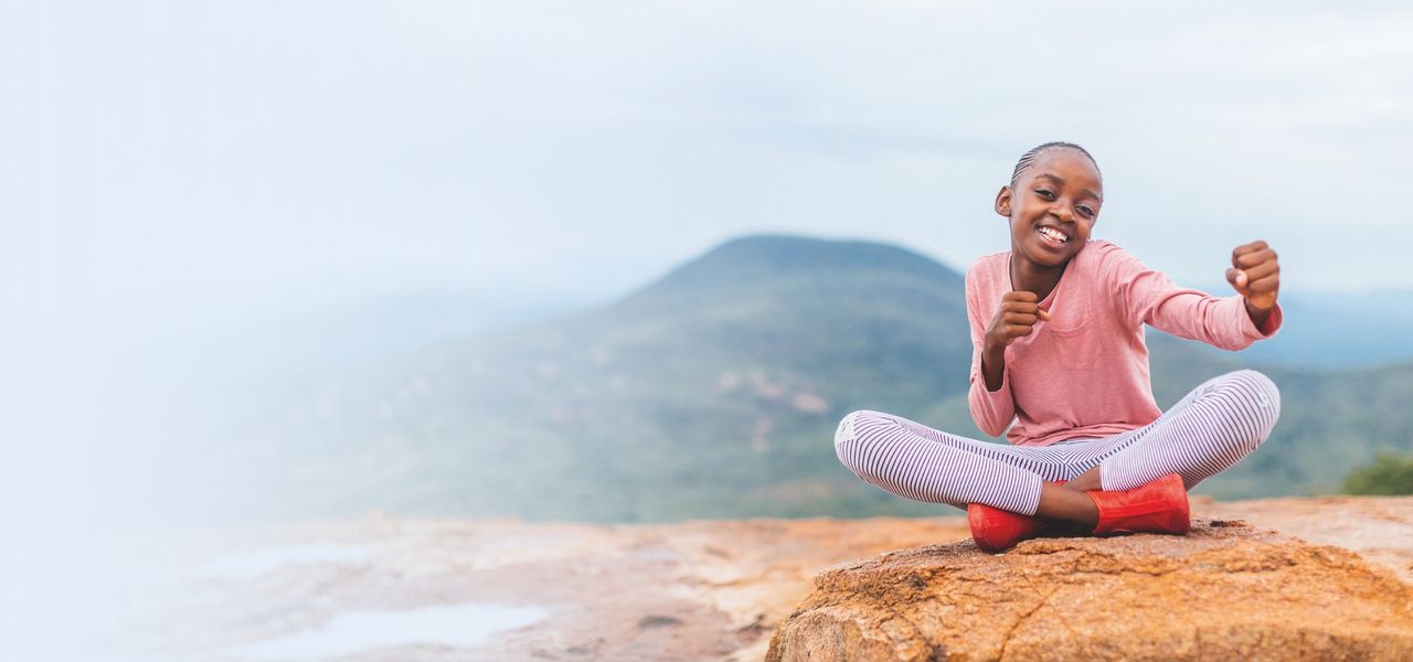 A young Octapharma patient in a pink shirt and striped leggings sits cross-legged on a rock, posing playfully with a scenic mountain landscape in the background.