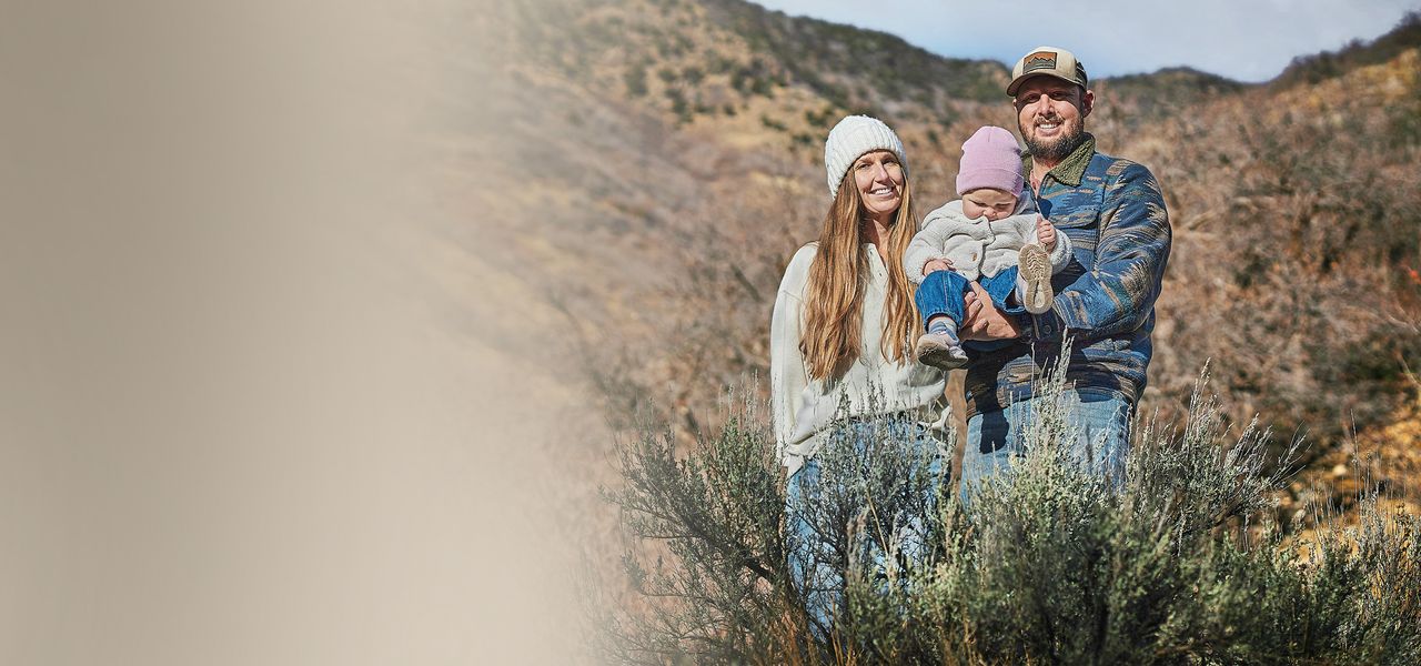 A smiling couple with a young child stand amidst brush with mountains in the background, on a sunny day - symbolizing sustainability and care for the environment.