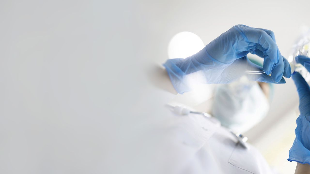 Medical professional wearing gloves and a mask examines an IV bag in a bright clinical environment