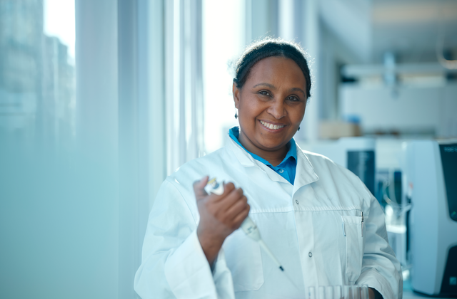 Smiling woman in a lab coat holding a test tube rack, standing in a brightly lit laboratory with equipment in the background.