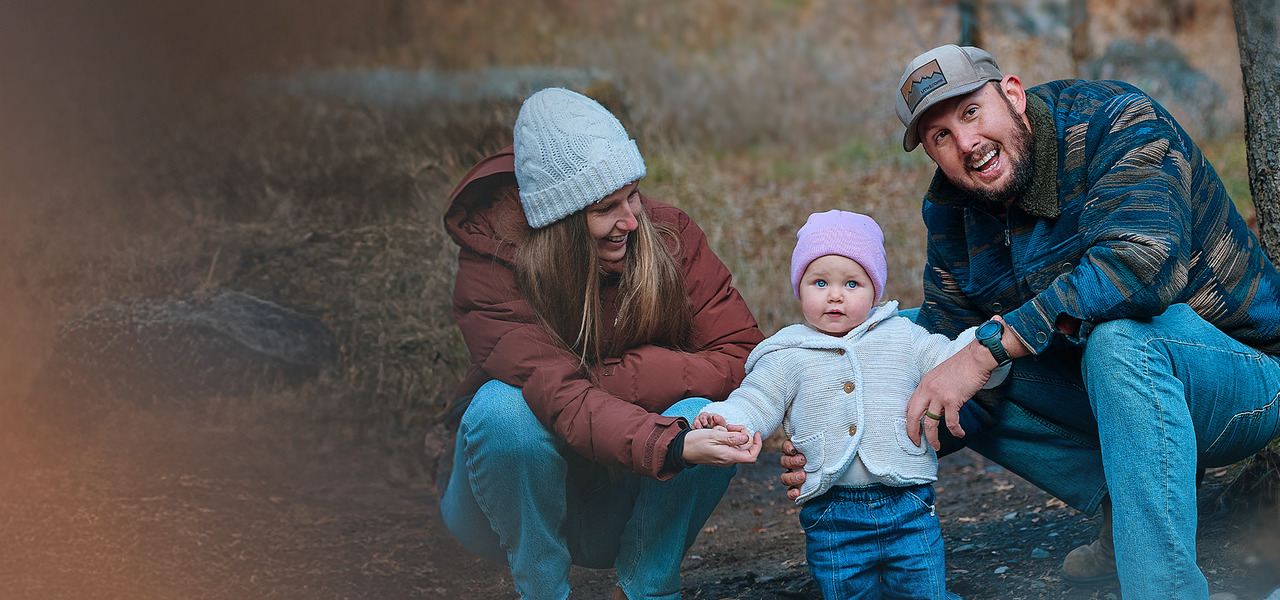 Eine Familie im Freien: Eine Frau und ein Mann hocken neben einem Baby mit rosa Mütze, alle warm eingepackt.