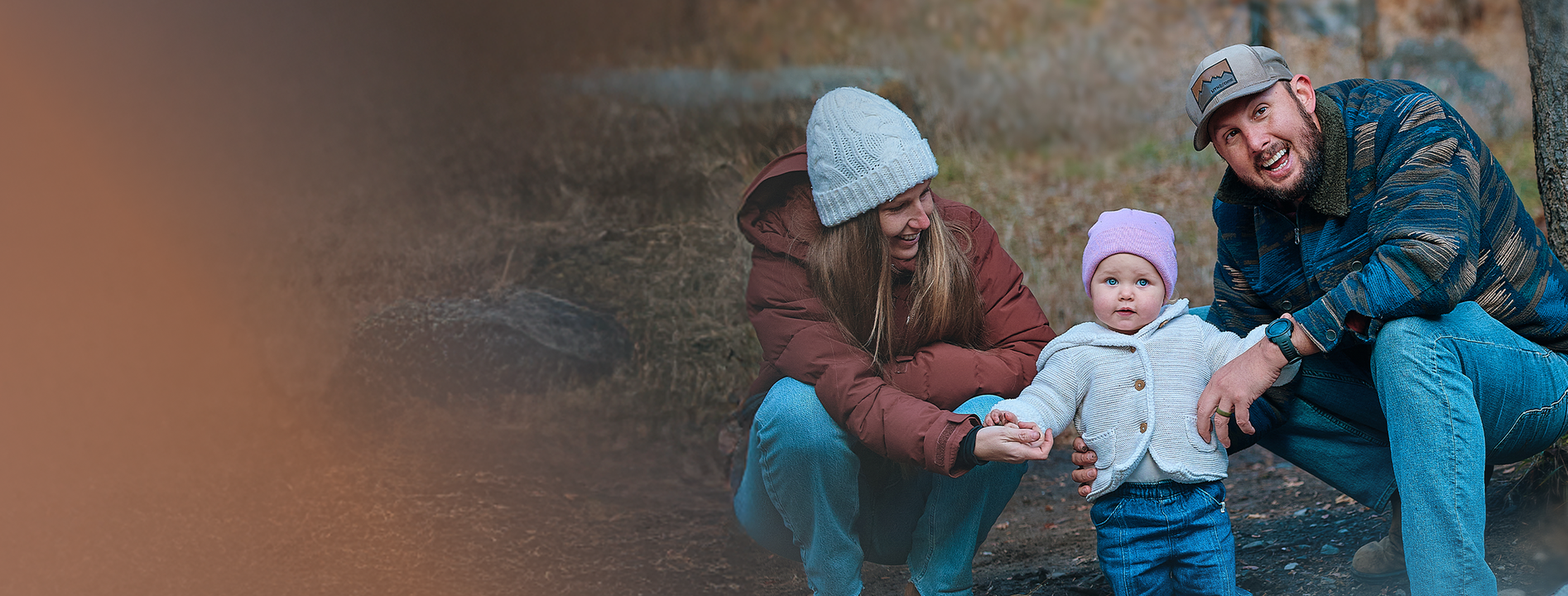 A family outdoors: a woman and man crouch beside a baby wearing a pink hat, all bundled up in warm clothing.