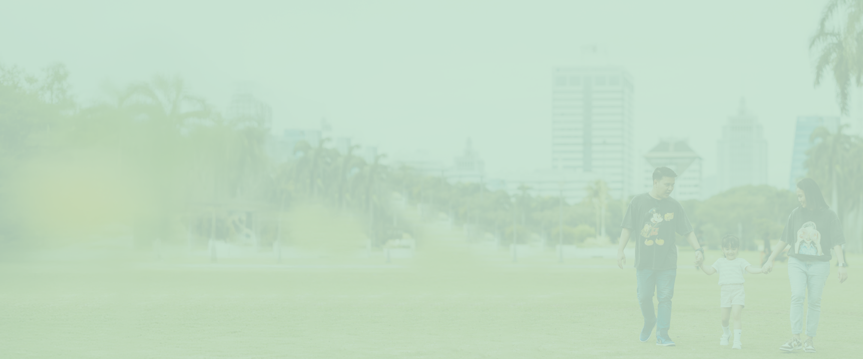 A family of three walks hand in hand across a grassy field in an urban park, with tall buildings and trees in the background.