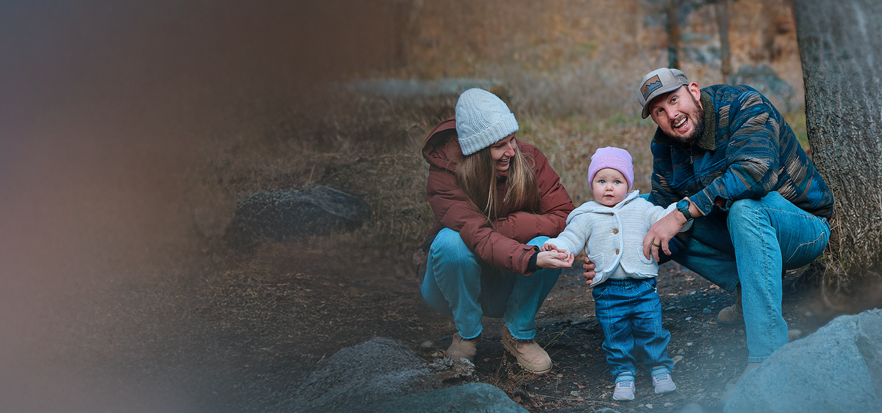 A family outdoors: a woman and man crouch beside a baby wearing a pink hat, all bundled up in warm clothing.