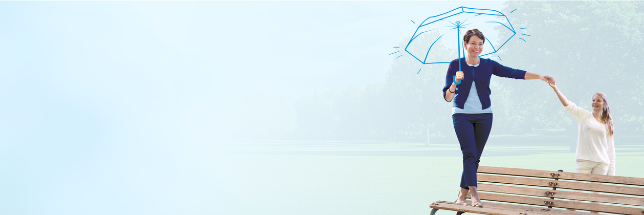 Woman balancing on a bench holding a blue-drawn umbrella with another woman supporting her in a park setting, clear blue sky background.