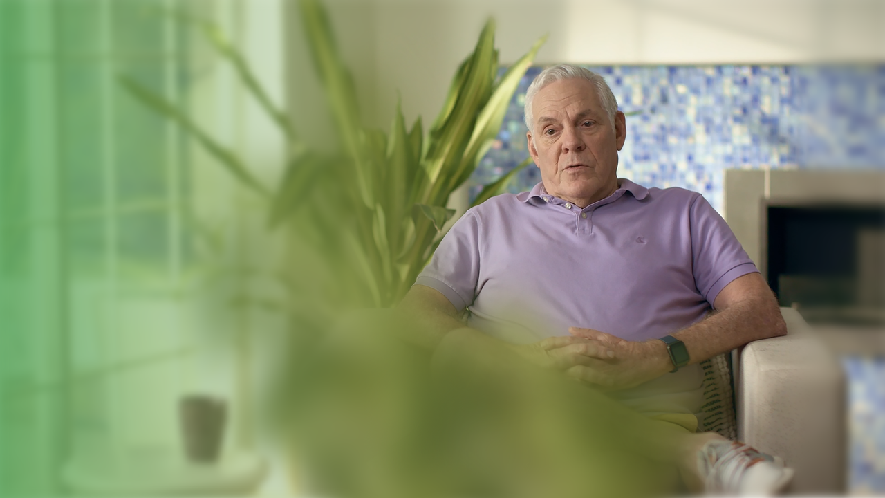 A man in a lavender polo shirt sits in a modern, light-filled room with a colorful tiled wall and green plants.