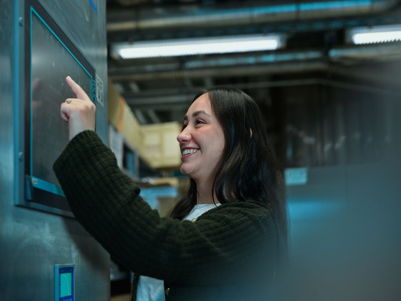 Smiling woman in a green sweater interacts with a touchscreen in an industrial setting.