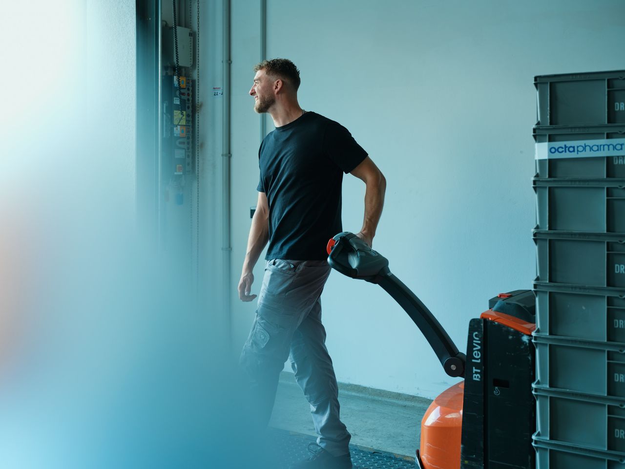 Man operating an electric pallet jack near stacked crates in a warehouse, wearing a black t-shirt and gray pants.