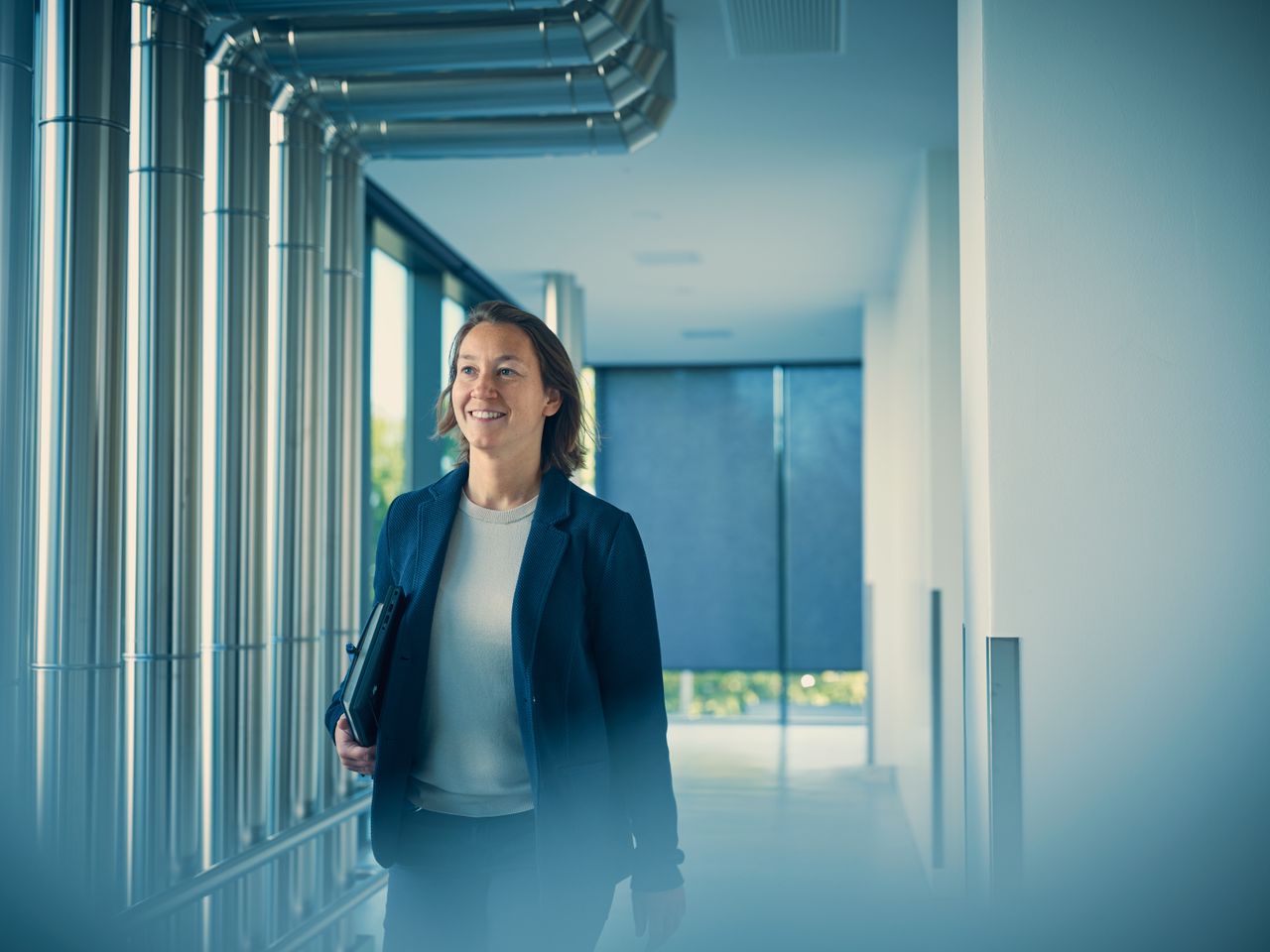 A person in a suit holds a folder, walking through a hallway with metal pipes and windows.