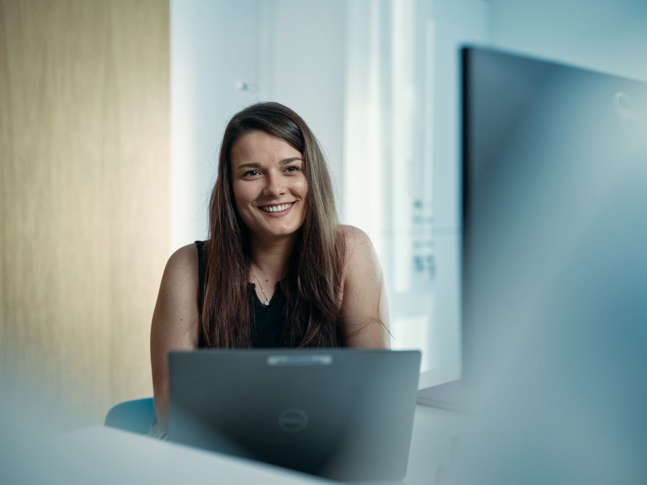 Une femme aux cheveux longs sourit, assise devant un ordinateur portable dans un bureau baigné de lumière.