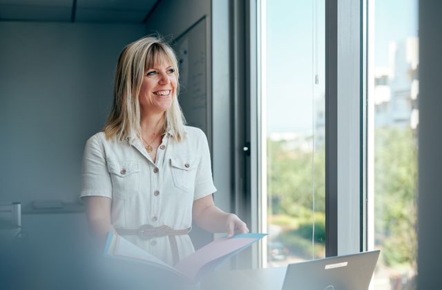 Une employée vêtue d'une robe blanche se tient près d'une fenêtre, un dossier à la main, et sourit. Un ordinateur portable est posé sur le bureau à proximité.