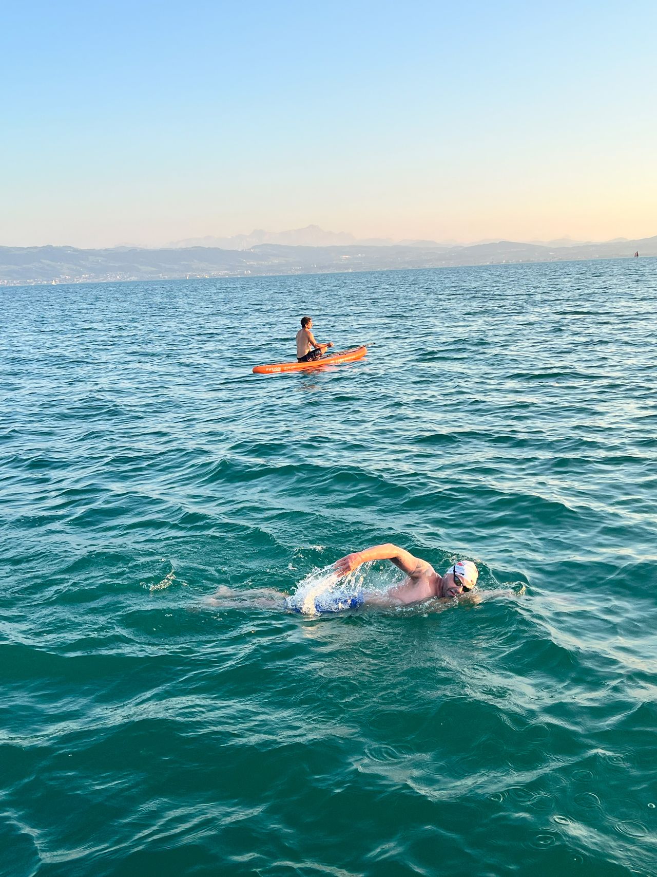 Neil swims in a lake, while another person in an orange kayak paddles nearby under a clear blue sky.