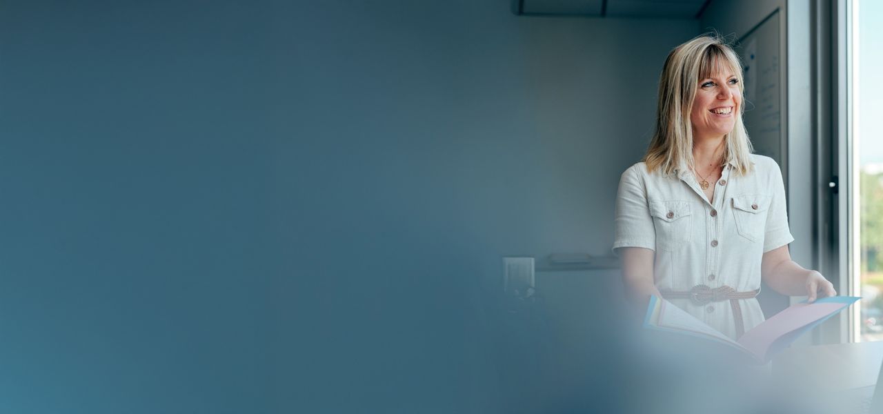 A female Octapharma employee in a white shirt stands by a window, holding a folder and smiling, with a blurred blue foreground.