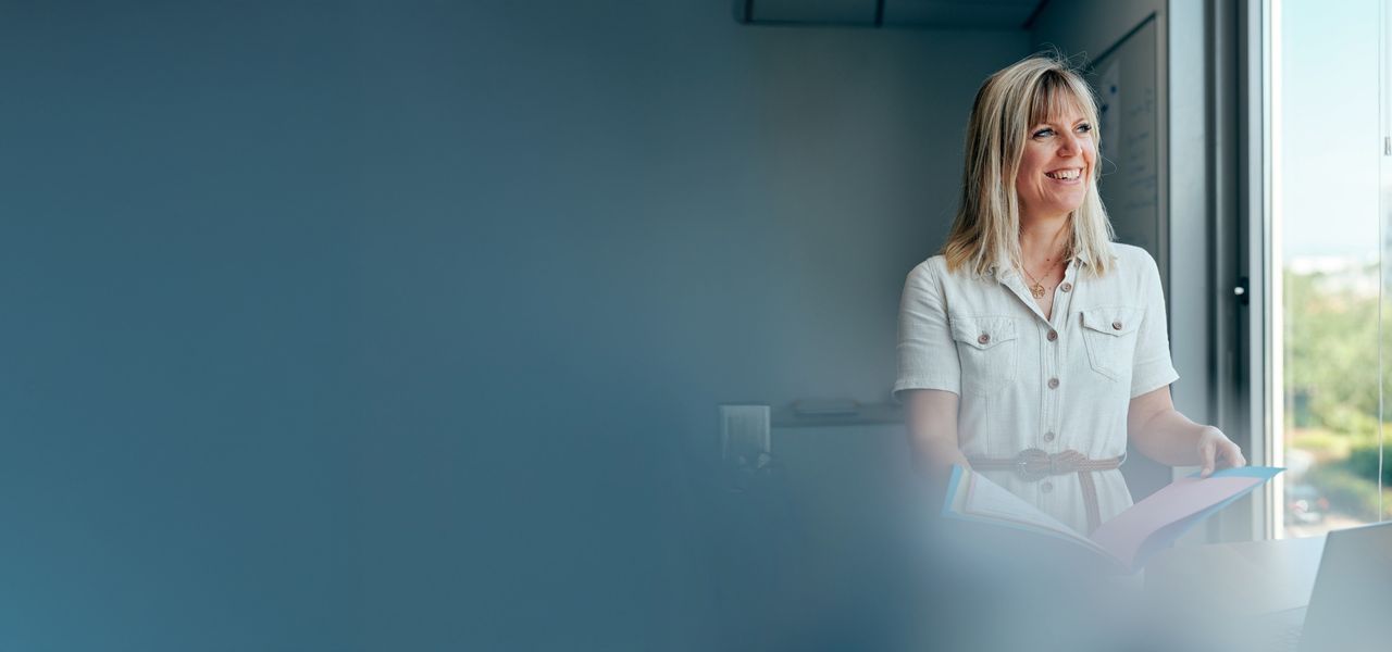 A female Octapharma employee in a white shirt stands by a window, holding a folder and smiling, with a blurred blue foreground.
