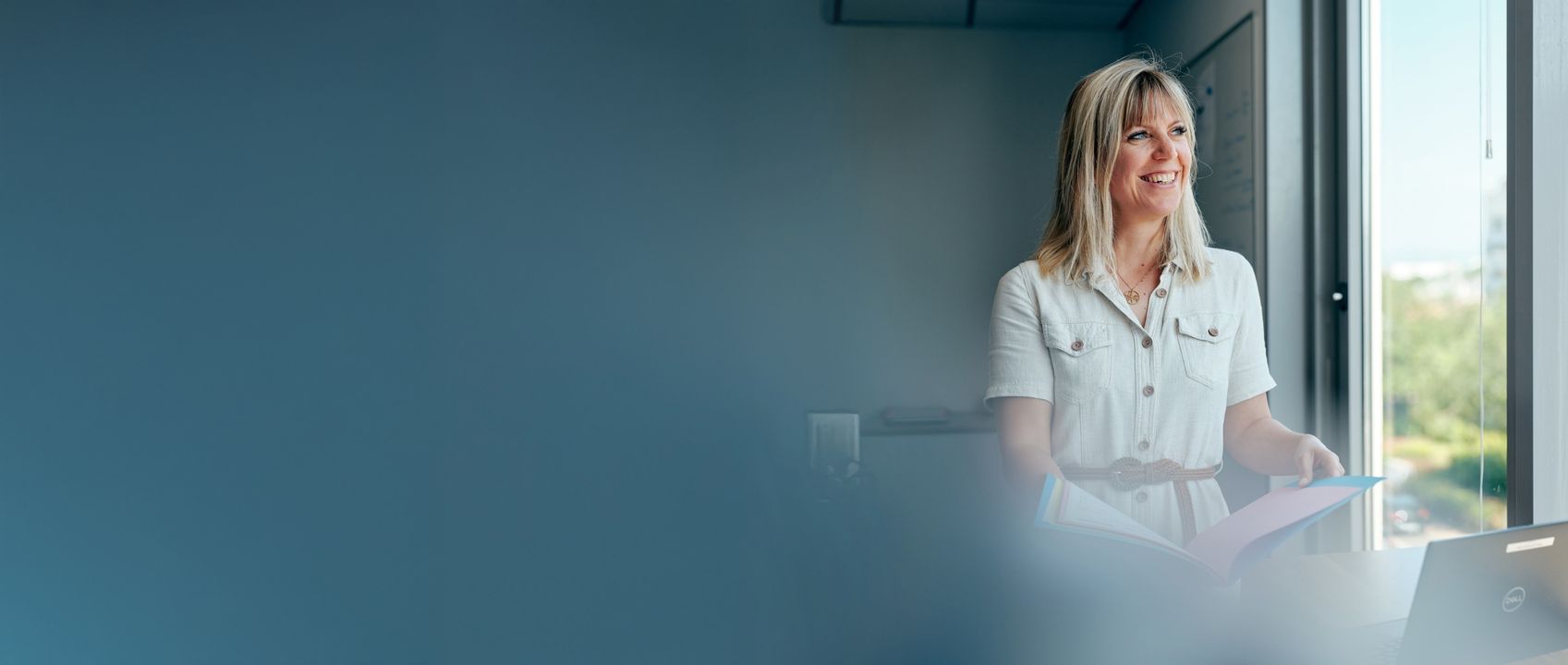 A female employee in a white shirt stands by a window, holding a folder and smiling, with a blurred blue foreground.