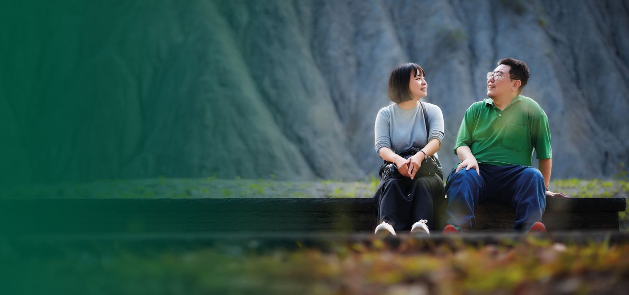 A man and woman sit on a bench outdoors, smiling at each other, with a steep rocky mountain backdrop.