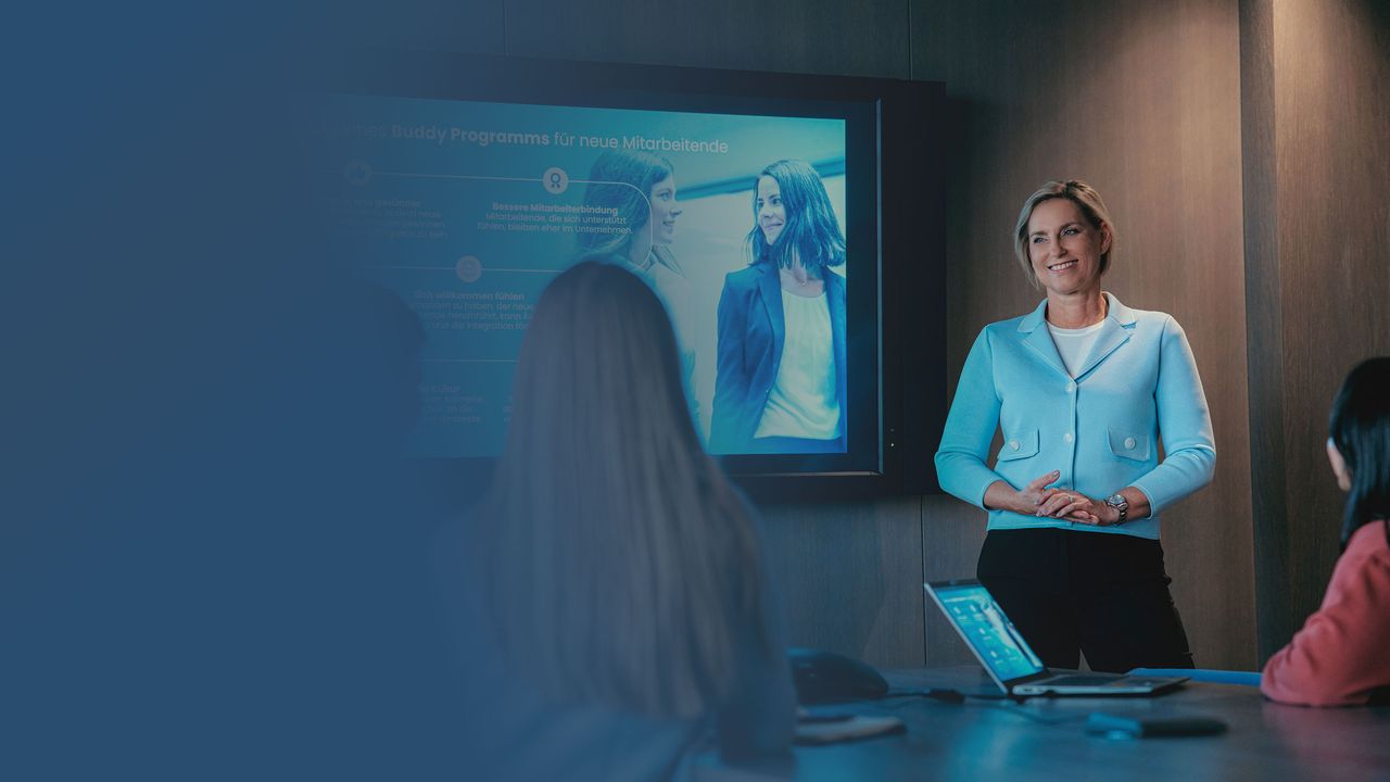 Bild von Gerda Brandt in einem Meeting mit 3 Kolleg:innen. Sie steht vor einem Screen und lächelt.