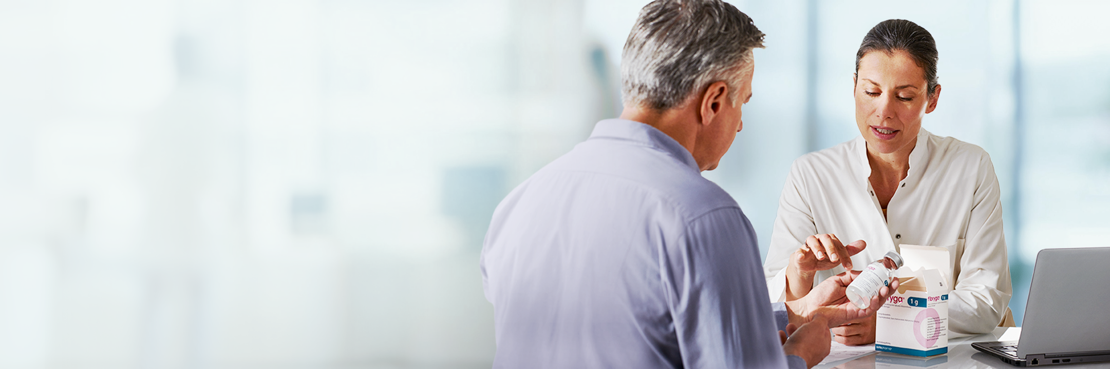 A pharmacist explains fibryga to an older male customer at a counter. A laptop is partially visible on the counter.