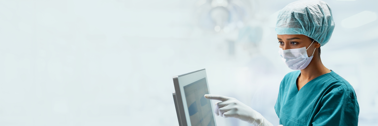 A medical professional in scrubs and a mask operates a touchscreen in a clinical environment.
