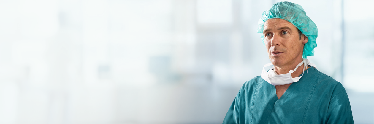 A healthcare professional in green scrubs, cap, and a surgical mask around the neck, stands in a bright medical setting.