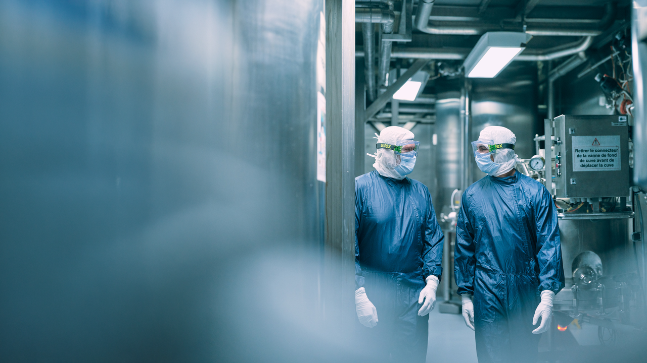 Deux personnes vêtues d'une tenue de protection, comprenant des lunettes et un masque, se déplacent dans un environnement industriel stérile.