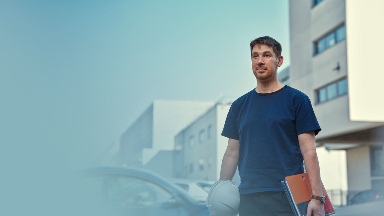 Un homme, Chef de Projet, est devant un chantier. Il porte un casque de chantier et un cahier dans ses mains. Il est débout, fier.