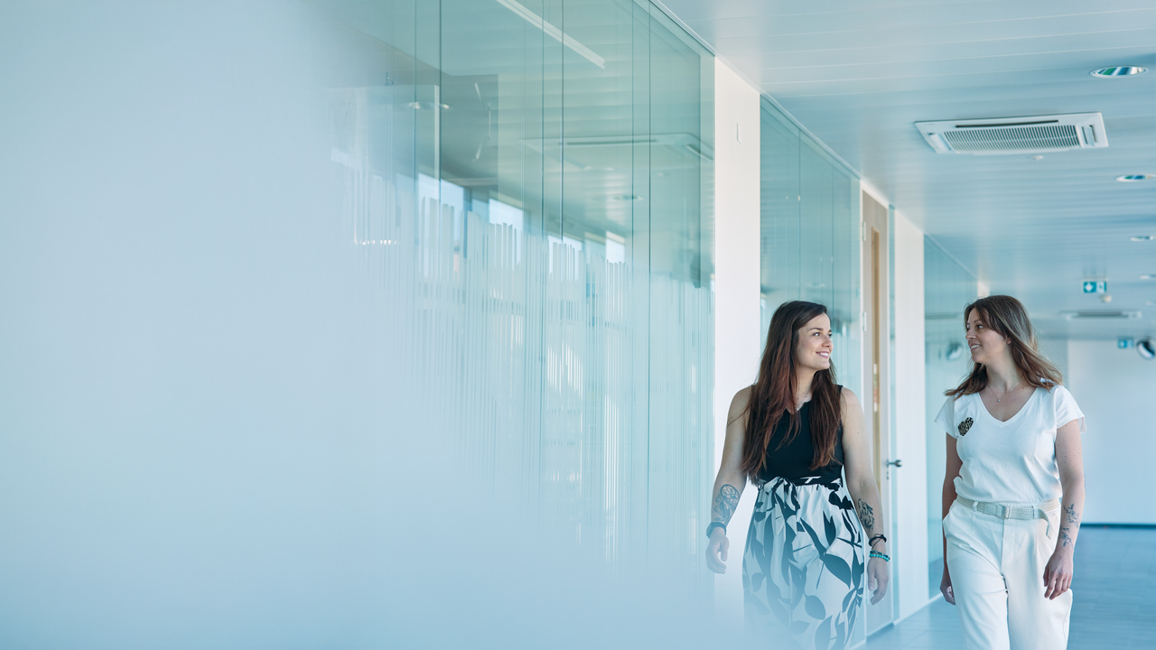 Deux femmes qui marchent en souriant dans un couloir lumineux et moderne, aux parois vitrées et baigné de lumière naturelle.