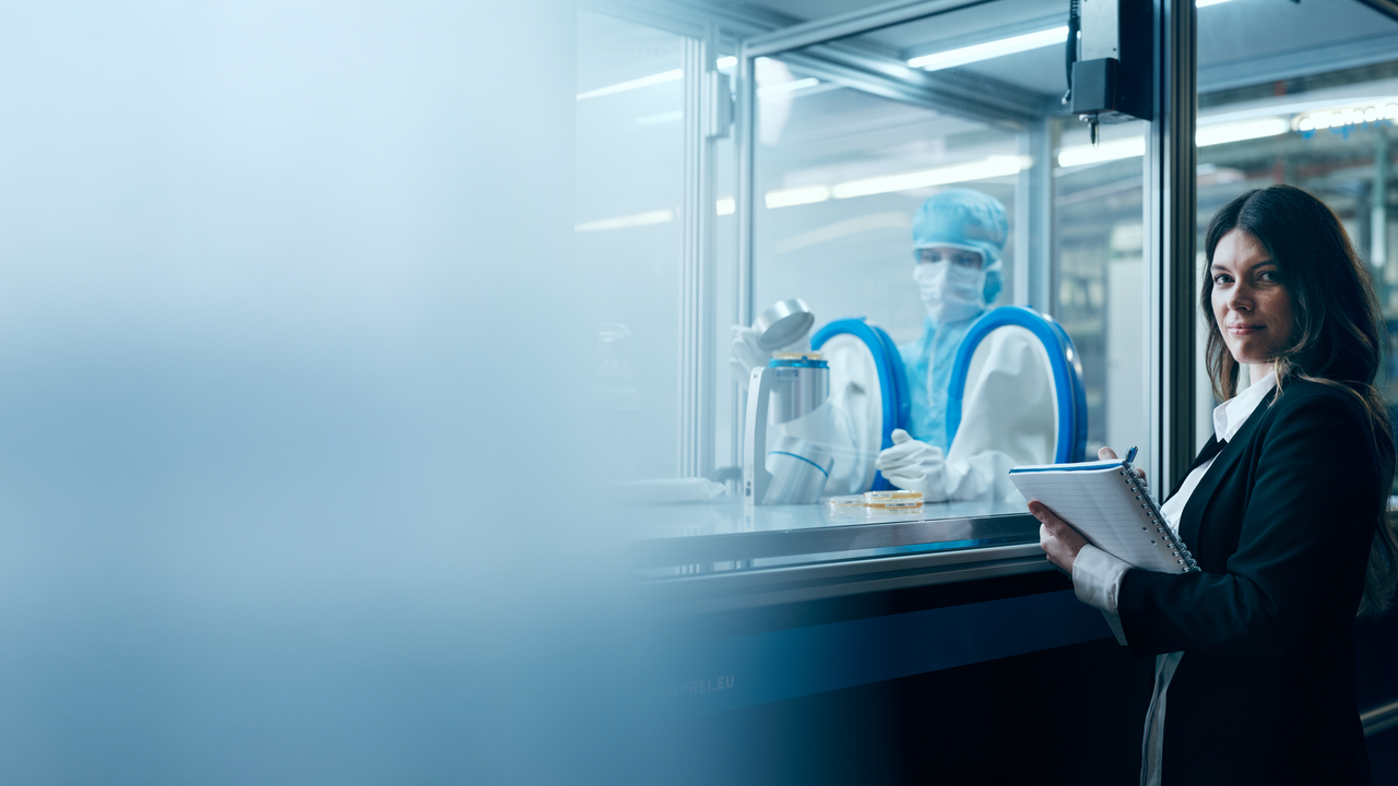 Bild von Viktoria Wiedemann auf dem sie vor einem Fenster steht hinter dem eine Testung durchgeführt wird. Viktoria blickt in die Kamera und hat einen Stift und Notizblock in ihrer Hand.