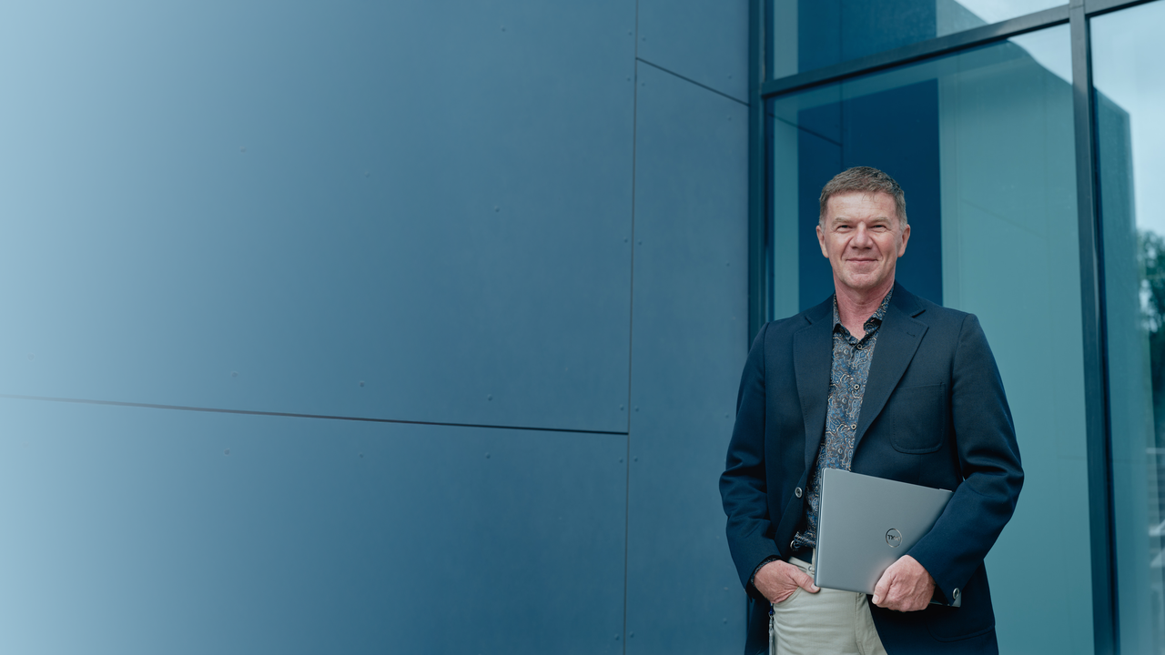 Bild auf dem Christian Korger vor einem Gebäude von Octapharma positioniert ist. Er trägt ein dunkles Sakko und einen Laptop unter seinem linken Arm.