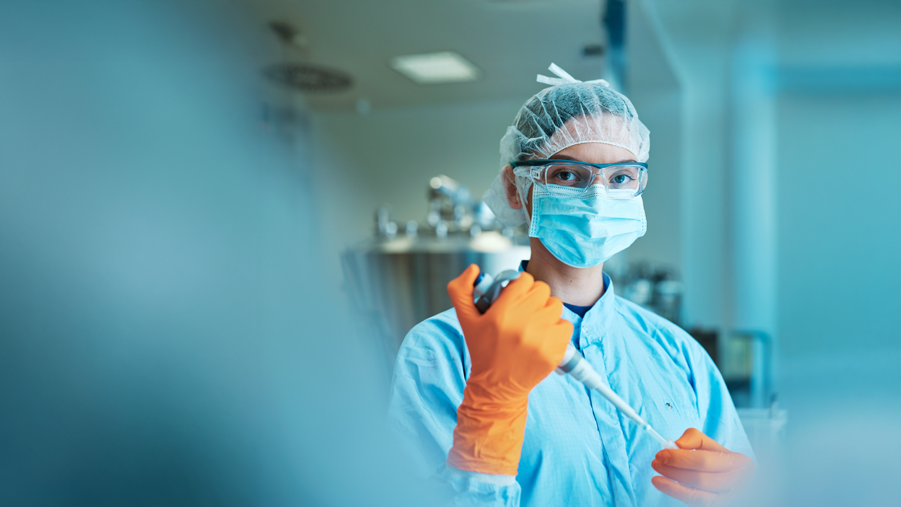 A lab technician wearing a mask, hairnet, and gloves holds a pipette in a laboratory setting..