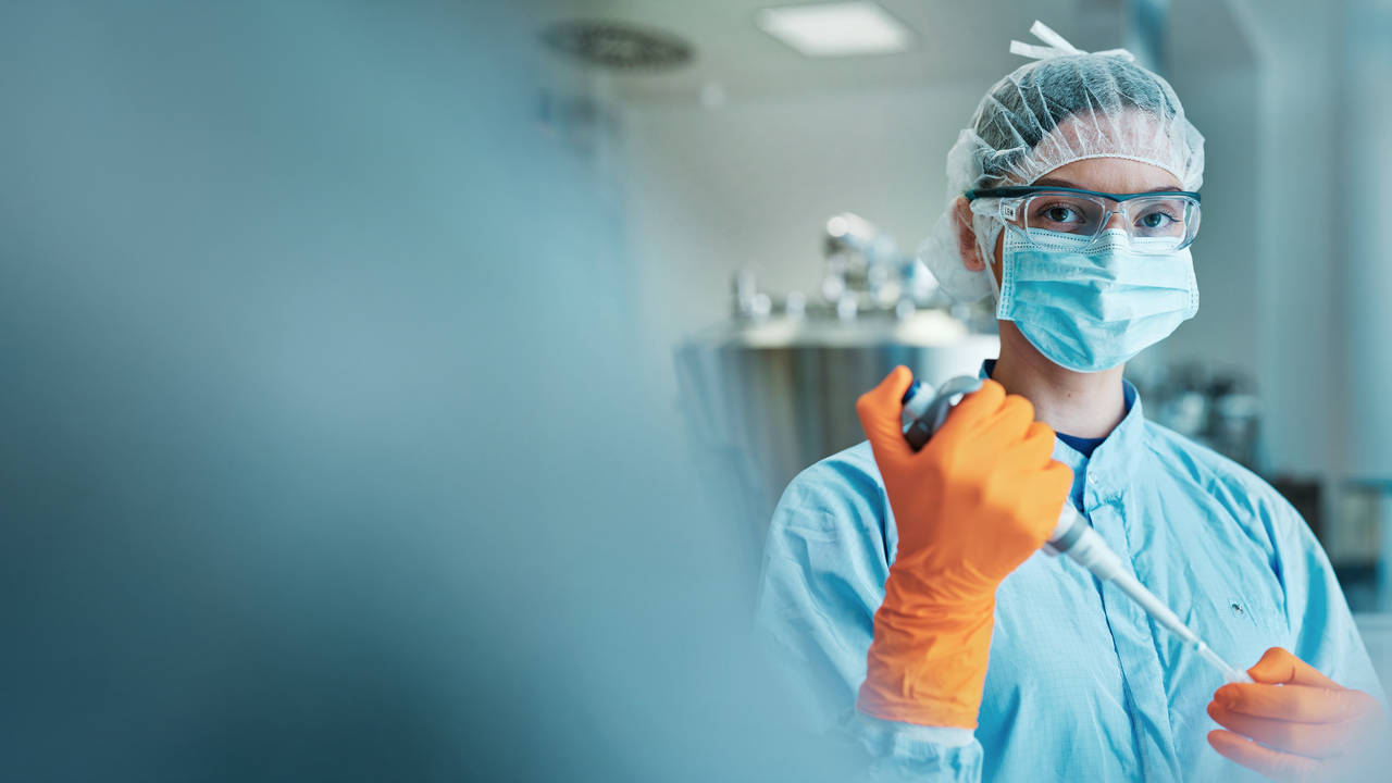 Ein Mitarbeiterin in Schutzkleidung und orangefarbenen Handschuhen hält eine Pipette in einer Laborumgebung und blickt direkt in die Kamera.