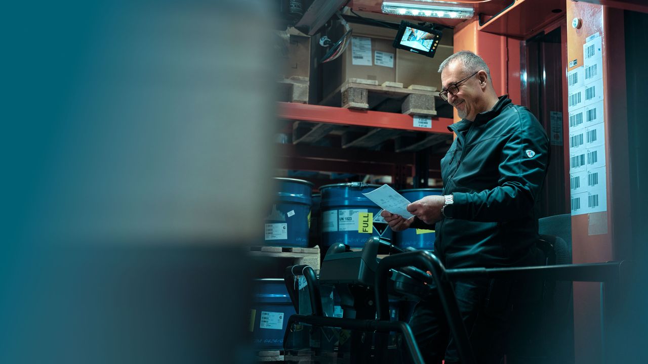 A men in a warehouse reads documents, surrounded by blue drums and shelves, with a focused expression, under industrial lighting.