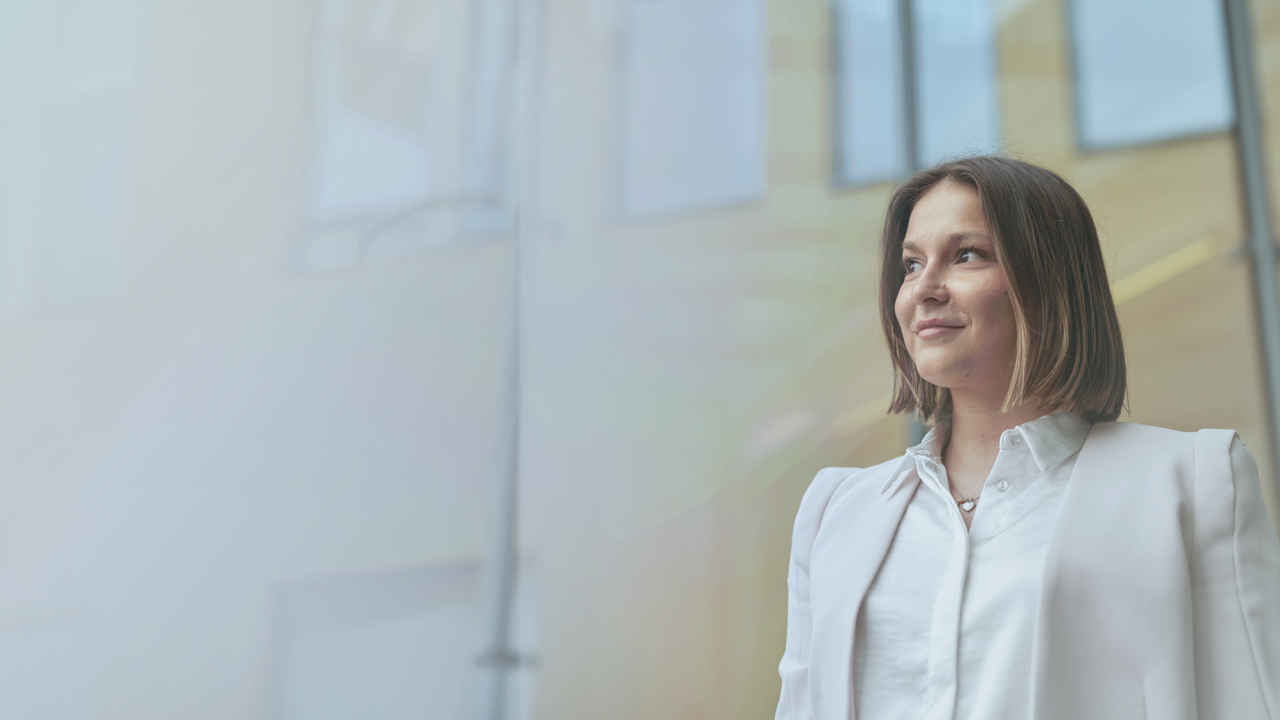 Bild auf dem Carina Niss vor einem Gebäude von Octapharma steht und nach links blickt. Carina trägt eine weiße Bluse und einen hellen Blazer.