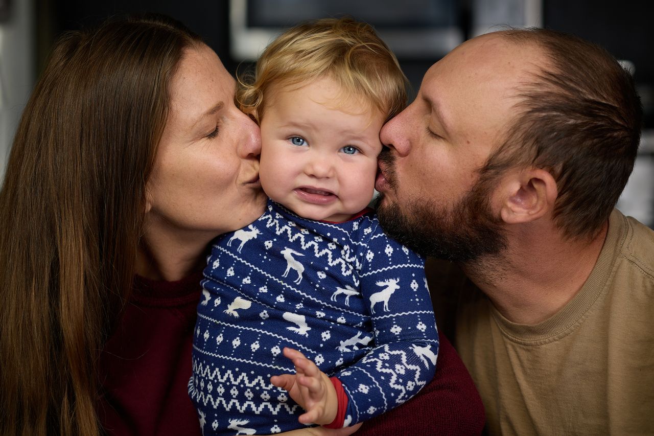 A child in a festive sweater is being kissed on both cheeks by his parents, one on each side, with a slightly amused expression.