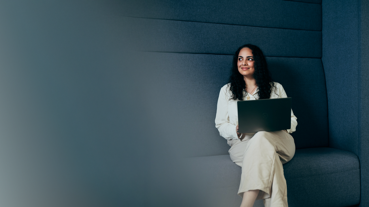 Bild von Periya Rafiei auf dem sie auf einem grauen Loungemöbel mit aufgeklapptem Laptop sitzt. Peryia blickt dabei nach links und lächelt.